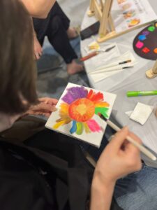 A person holds a canvas and paints a colorful, abstract flower using bright colors—an inspiring scene perfect for team building, with painting supplies, brushes, and paint visible on the table nearby.