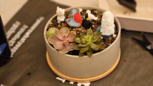 A small gray planter with succulents, decorative pebbles, and miniature figurines—including a penguin, a white animal, and a blue pond—sits on a round wooden tray, perfect for creative team building activities or thoughtful team building gifts.