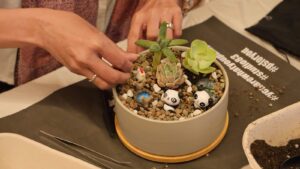 A person arranges small succulents and decorative figurines, including pandas, in a round pot filled with pebbles—an engaging Team Building activity. Gardening tools and soil are nearby on a table.
